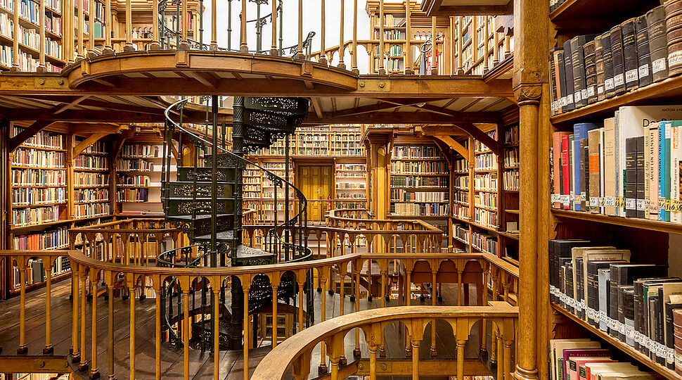 Viele hölzerne Bücherregale in verschiedenen Ebenen, auch eine gewundene Treppe ist zu sehen. Bibliotheken_150720-93-000176_web.jpg